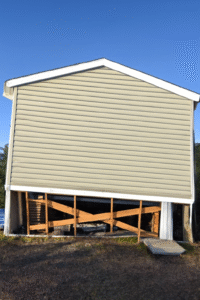 Mobile home leaning to the right with exposed support blocks and damaged skirting before releveling